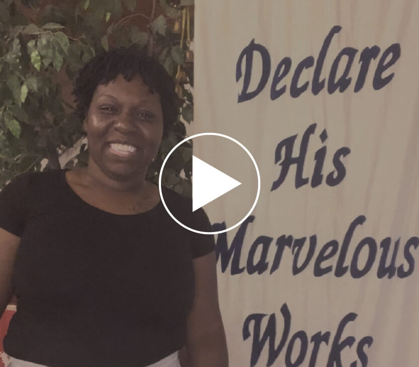 Monique standing in front of a banner that says Declare His marvelous works and a video play button